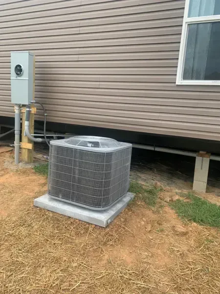 An outdoor HVAC condenser unit sits on a concrete pad next to a beige vinyl-sided building with an electrical meter box.