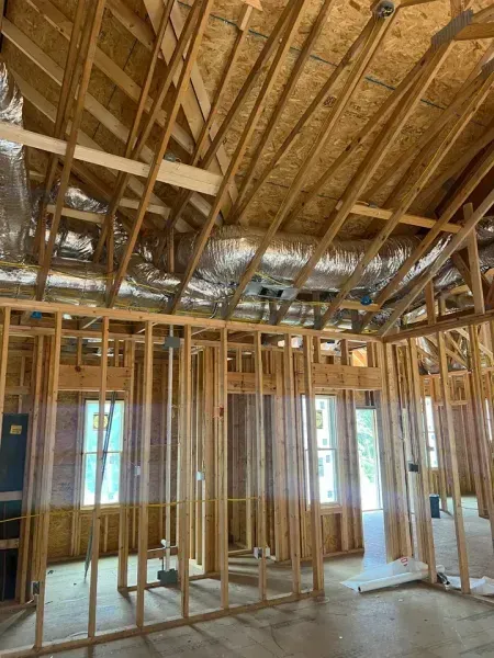 An interior view of a room under construction, showing wooden wall framing, roof trusses, and exposed silver ductwork.