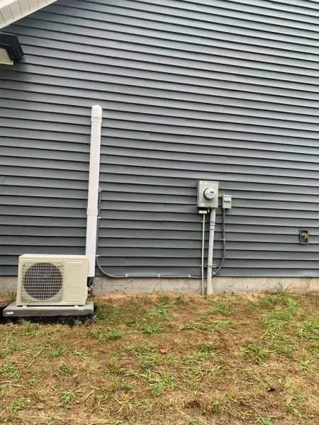 A small, beige outdoor HVAC unit stands beside a white vent pipe against the dark gray siding of a house exterior.