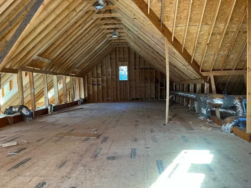 An unfinished attic with exposed wooden rafters, plywood subflooring, a central window, and exposed ductwork.