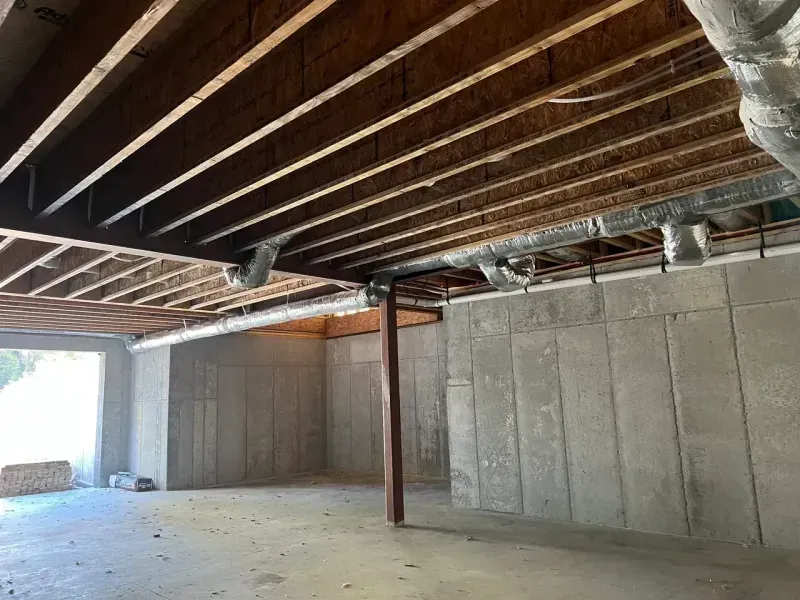 An unfinished basement with exposed wooden joists, concrete walls, a steel support column, and metal HVAC ductwork.