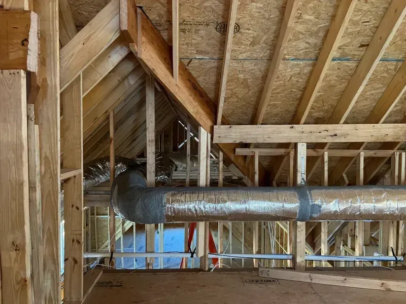 Construction site showing wood framing of a house roof, exposed rafters, and metallic HVAC ductwork in the attic area.