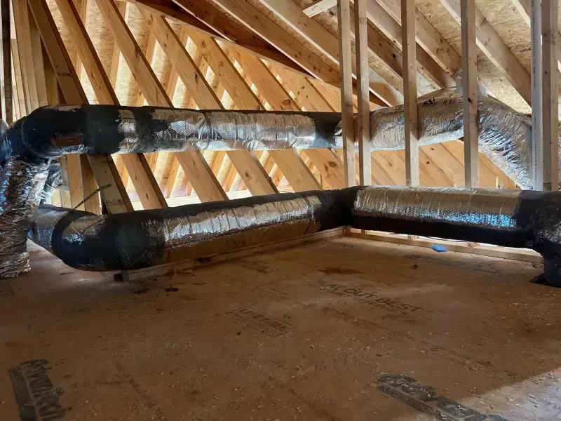 Insulated HVAC ductwork runs along the floor of an unfinished wooden attic space with visible roof rafters.
