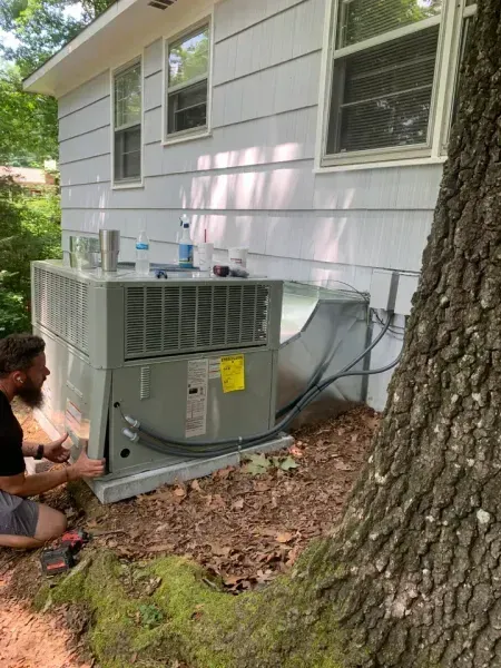 A technician in a black shirt works on a light gray outdoor HVAC unit next to a house exterior and a large tree.