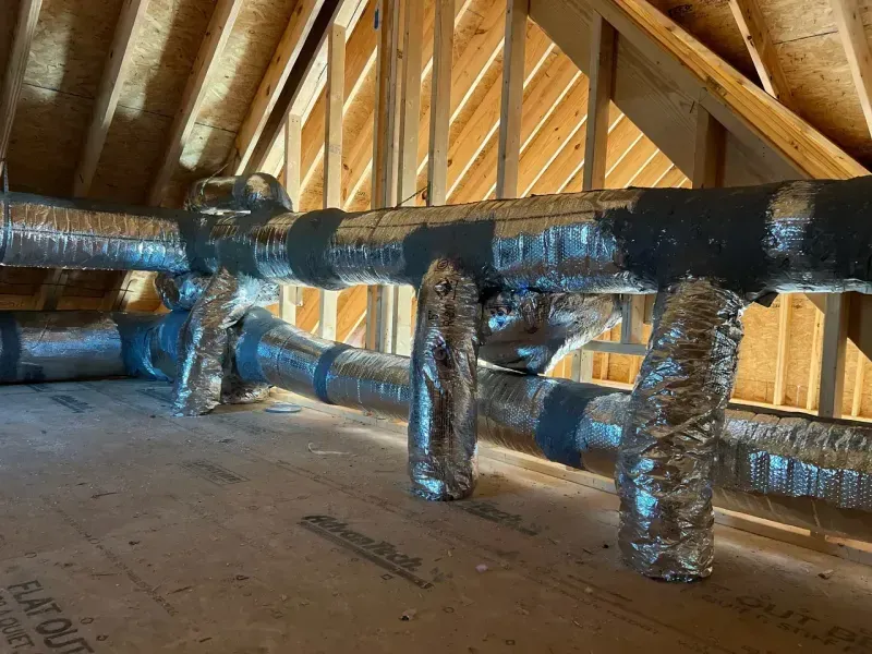 Silver, foil-wrapped HVAC air ducts run horizontally and vertically across an unfinished wooden attic floor.