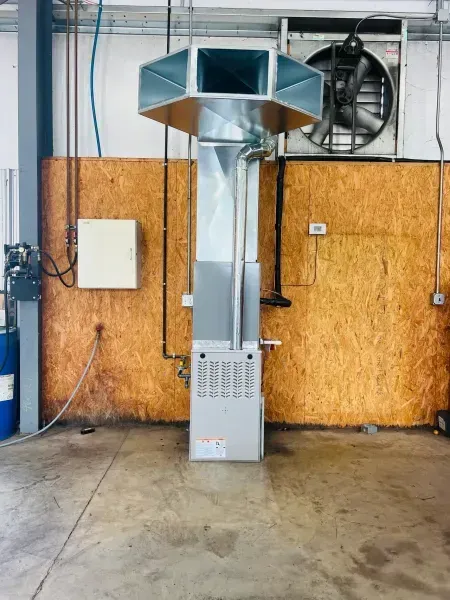 A furnace unit stands against a wooden wall, with a metal duct system extending upward toward a mounted wall exhaust fan.