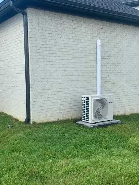 An outdoor HVAC condenser unit sits on a concrete pad next to a white brick wall, connected to a vertical pipe.