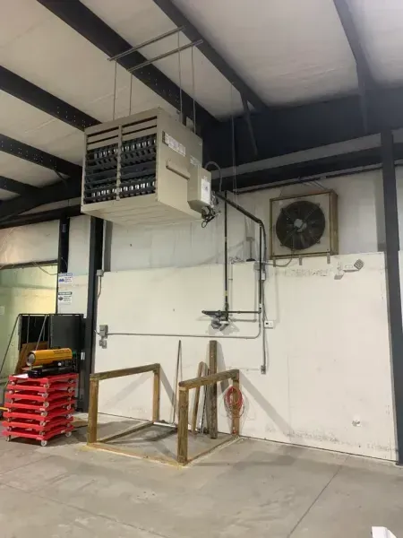 A large beige industrial heater suspended from the ceiling above a wall-mounted fan in a warehouse space.