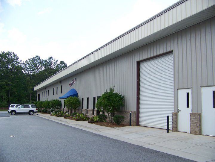 Exterior of a light-colored industrial building with a rollup door, a car parked, and landscaping.
