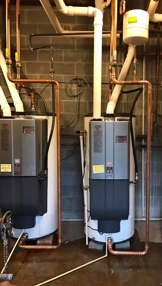 Two water heaters with copper pipes in a utility room against cinder block walls.