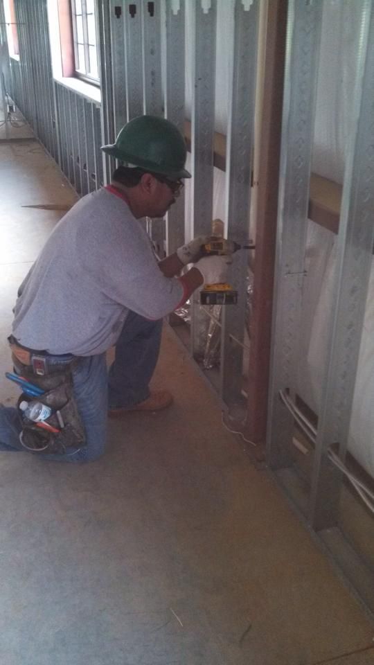Construction worker kneels, working on metal framing. He wears a green hard hat, glasses, and jeans, with a tool belt.