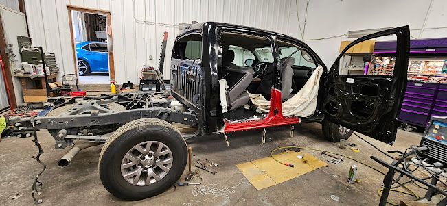 Truck frame in a repair shop with its doors open. The blue car is in the background.