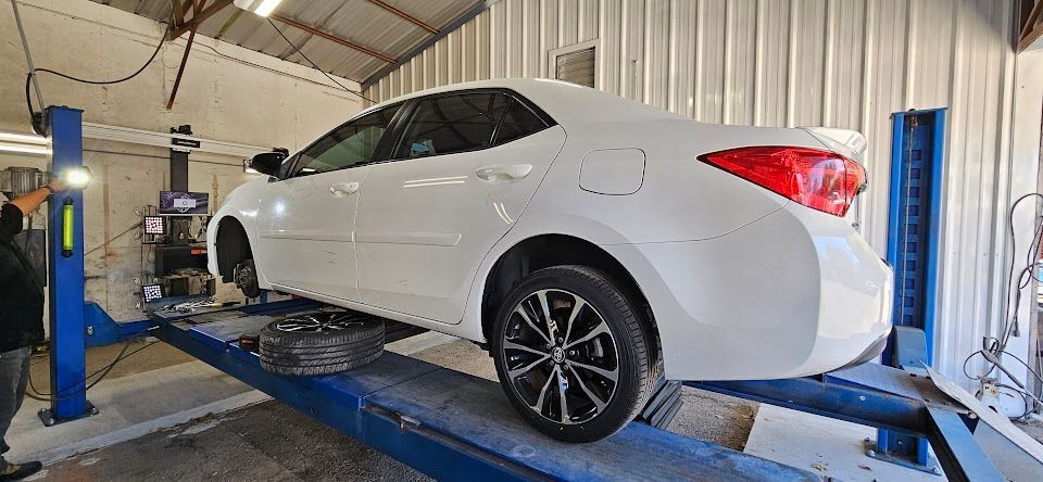 A white car on a lift in a garage. A person stands nearby. One tire is off.
