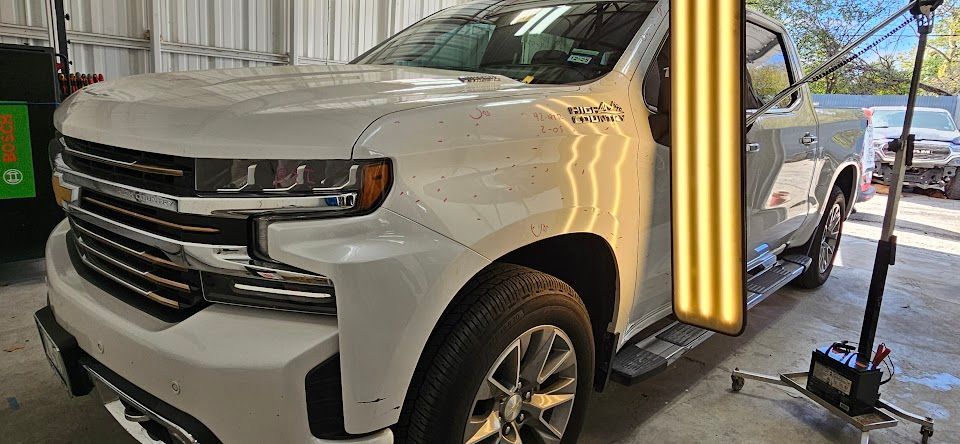 A white Chevrolet pickup truck in a repair shop with a dent. A technician is working on the dent.