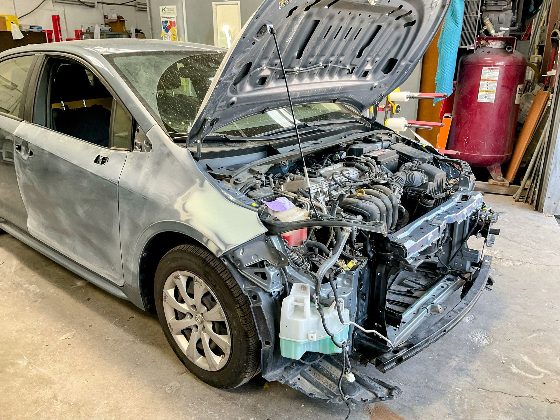 Car being repaired in a garage; hood open, front end disassembled, gray body, red air compressor in background.