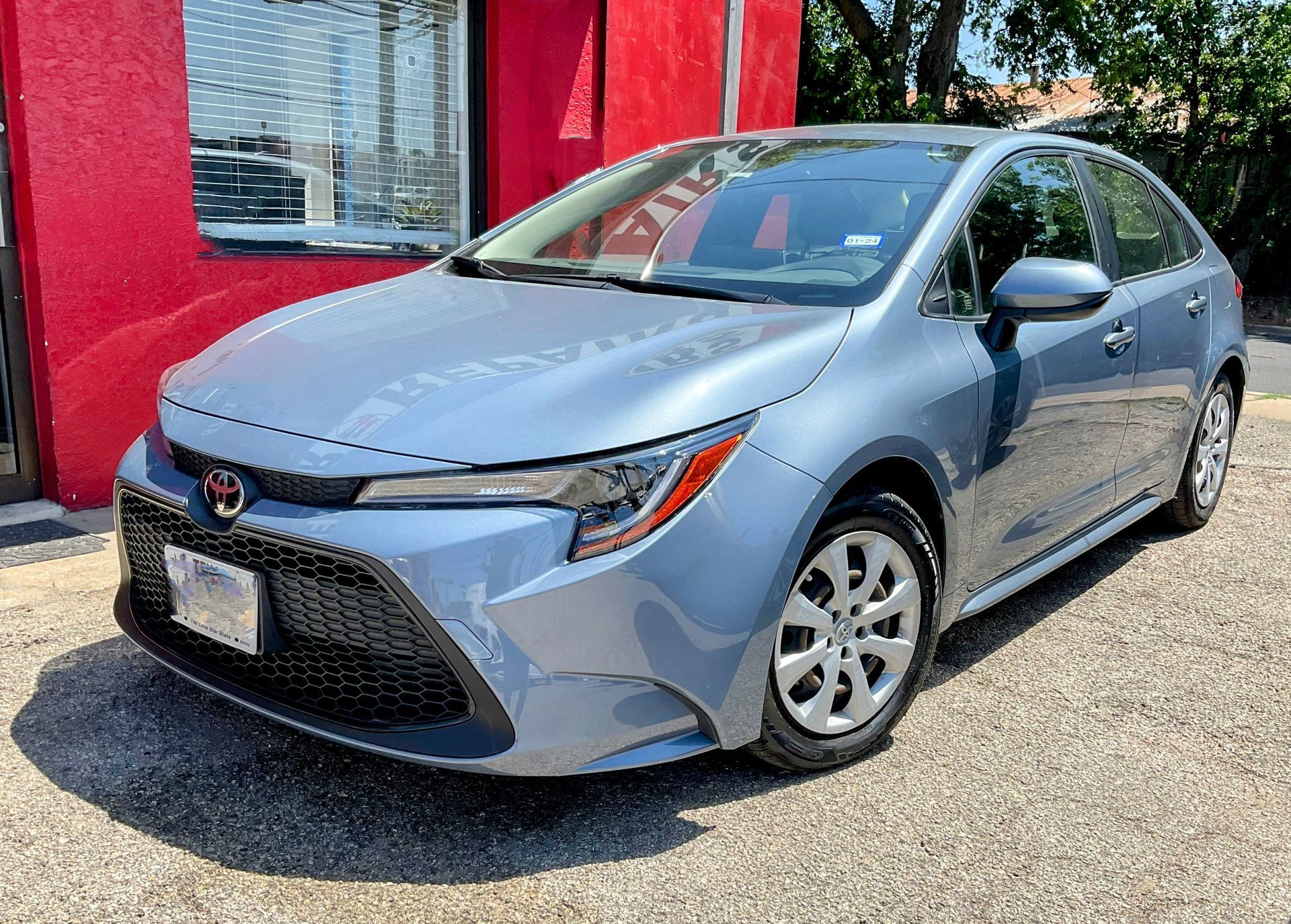 Blue Toyota Corolla hatchback parked in front of a red building on a sunny day.