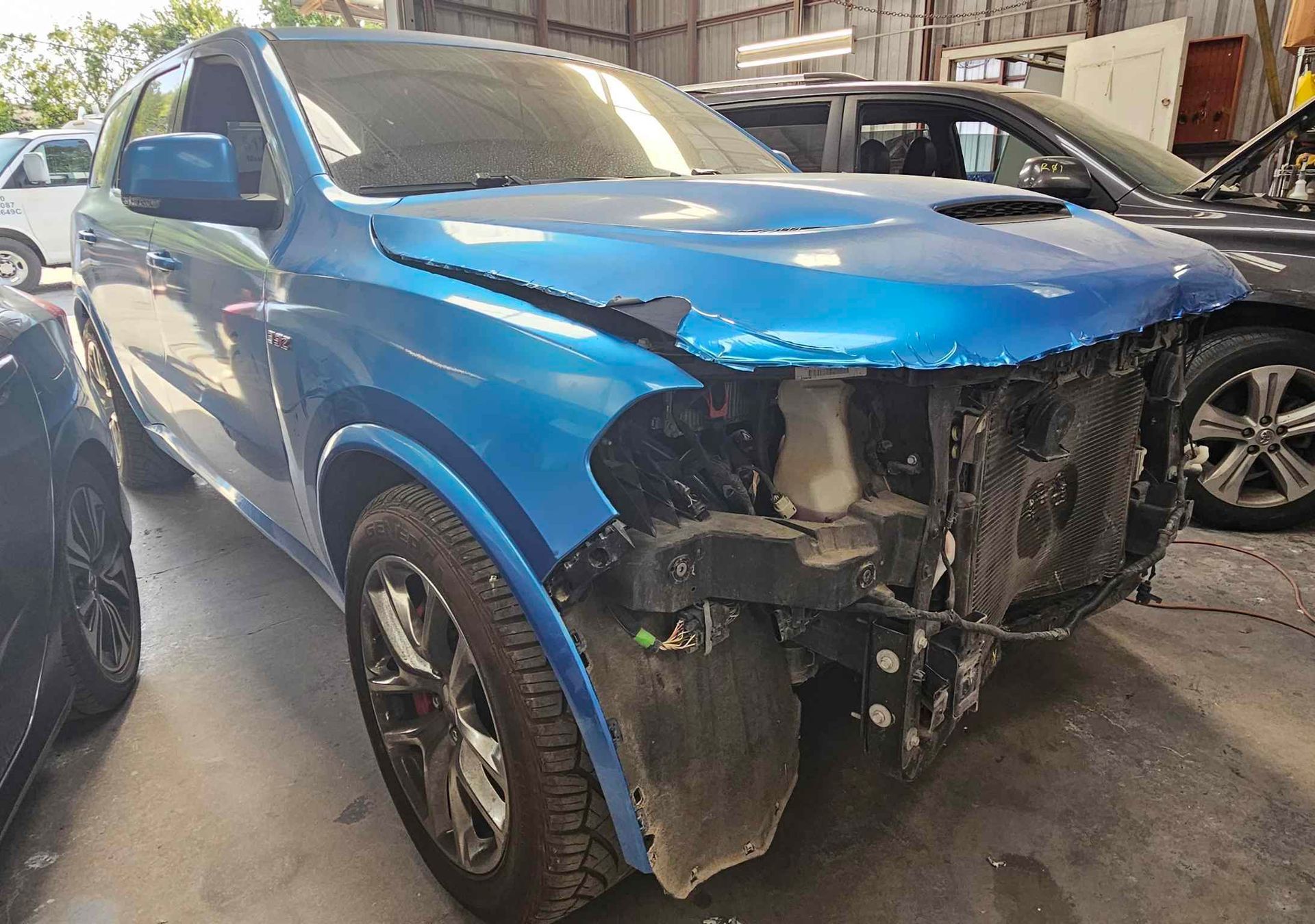 Damaged blue SUV in a repair shop, front end collision visible.