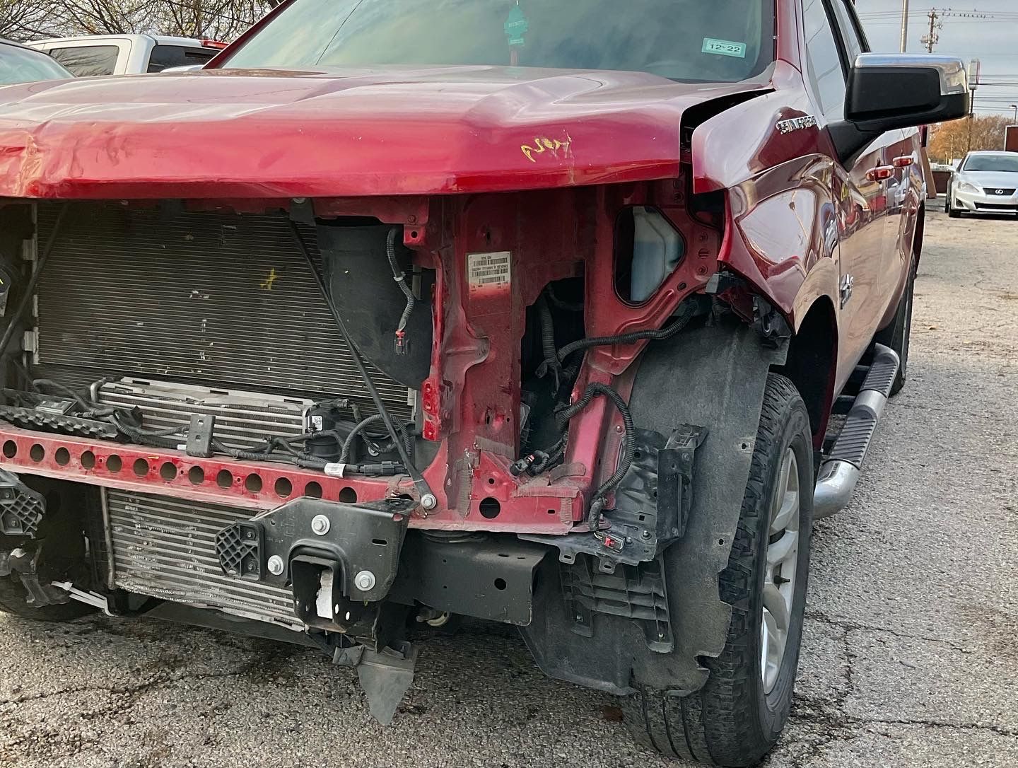 Red truck with front-end damage, missing bumper and headlight, in a junkyard.