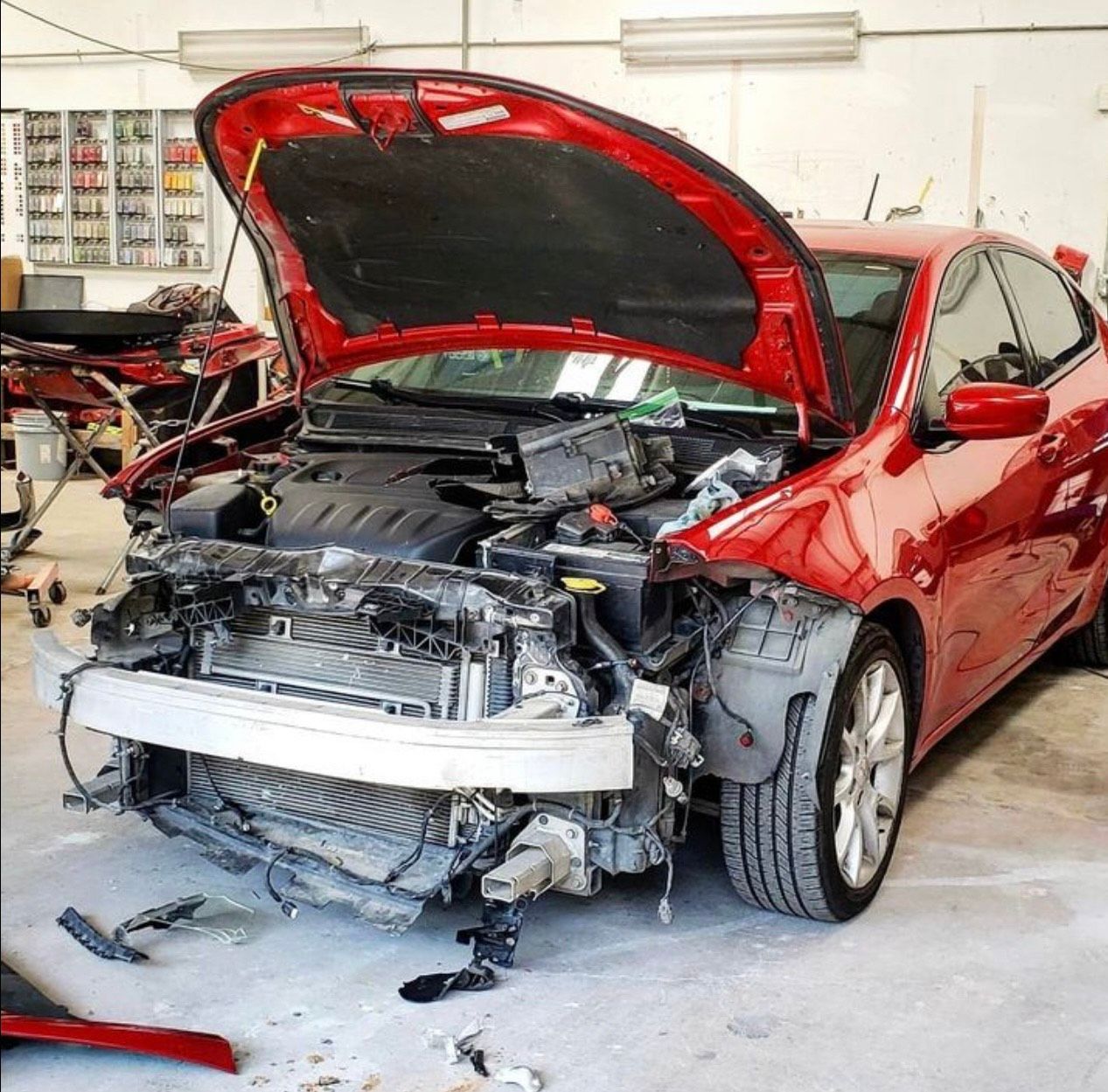 Red car with extensive front-end damage; hood open, parts scattered in a repair shop.