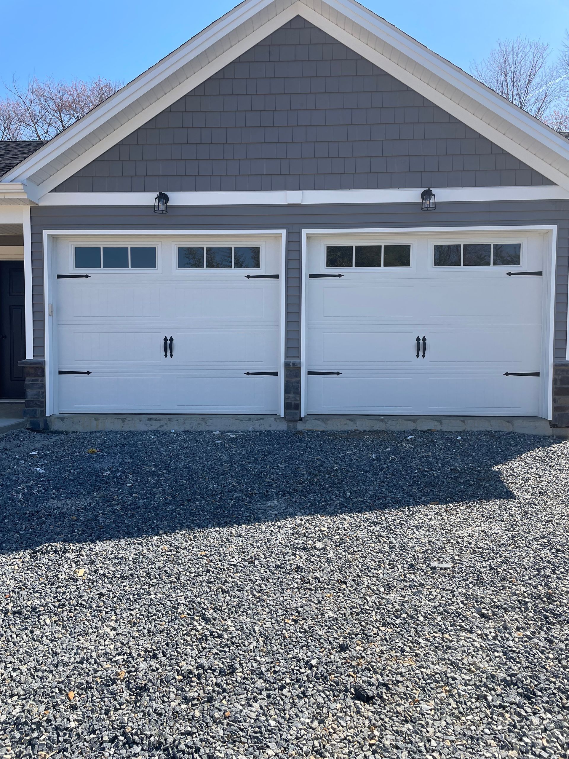 Two white garage doors on a gray house with a gravel driveway in front.