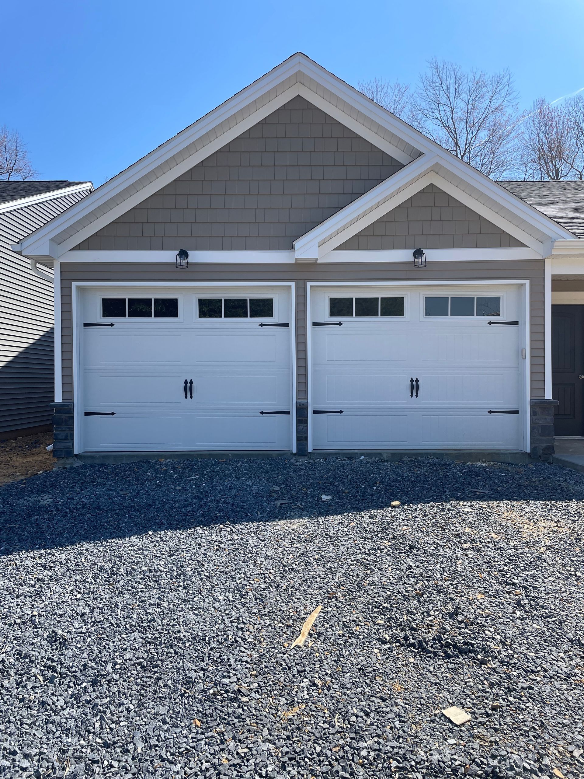 Two-car garage with white doors and gray siding, viewed from a gravel driveway under a clear blue sky