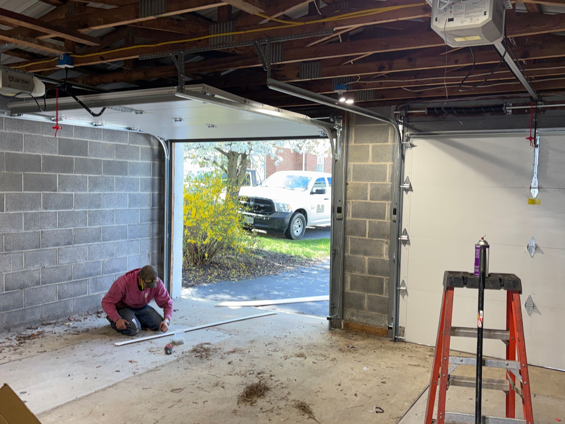 Person kneeling in a garage, working near an open door with a truck outside and a ladder inside.