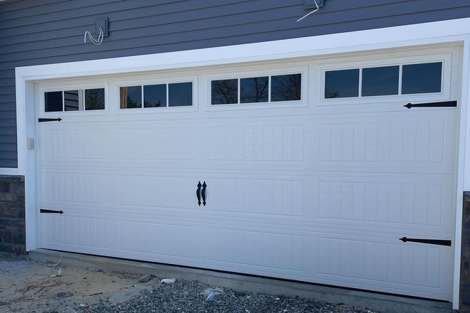 A white, paneled residential garage door with top windows and decorative black hardware against a gray siding wall.