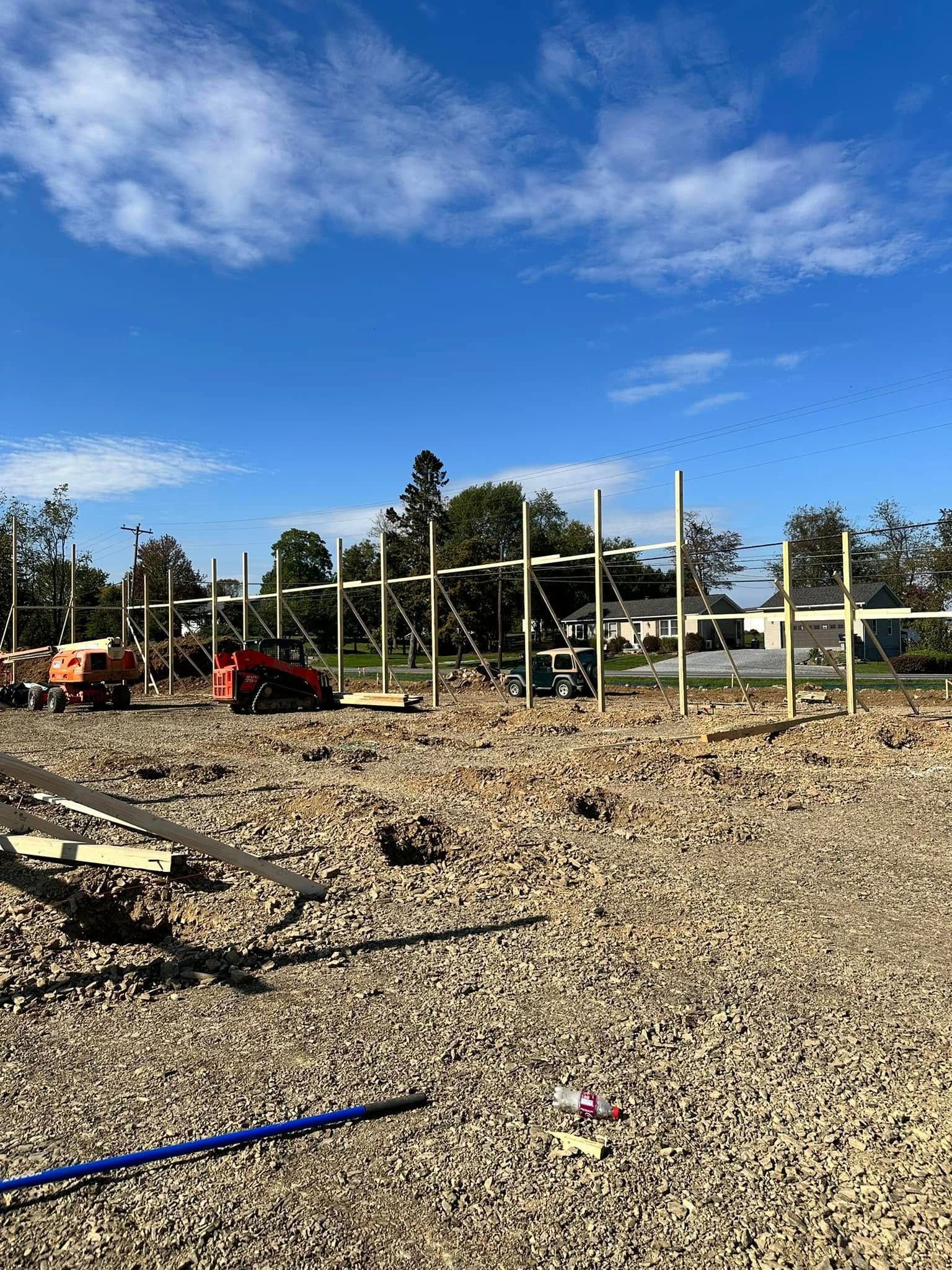Construction site showing a series of vertical wooden posts and framing under a blue sky with scattered gravel.