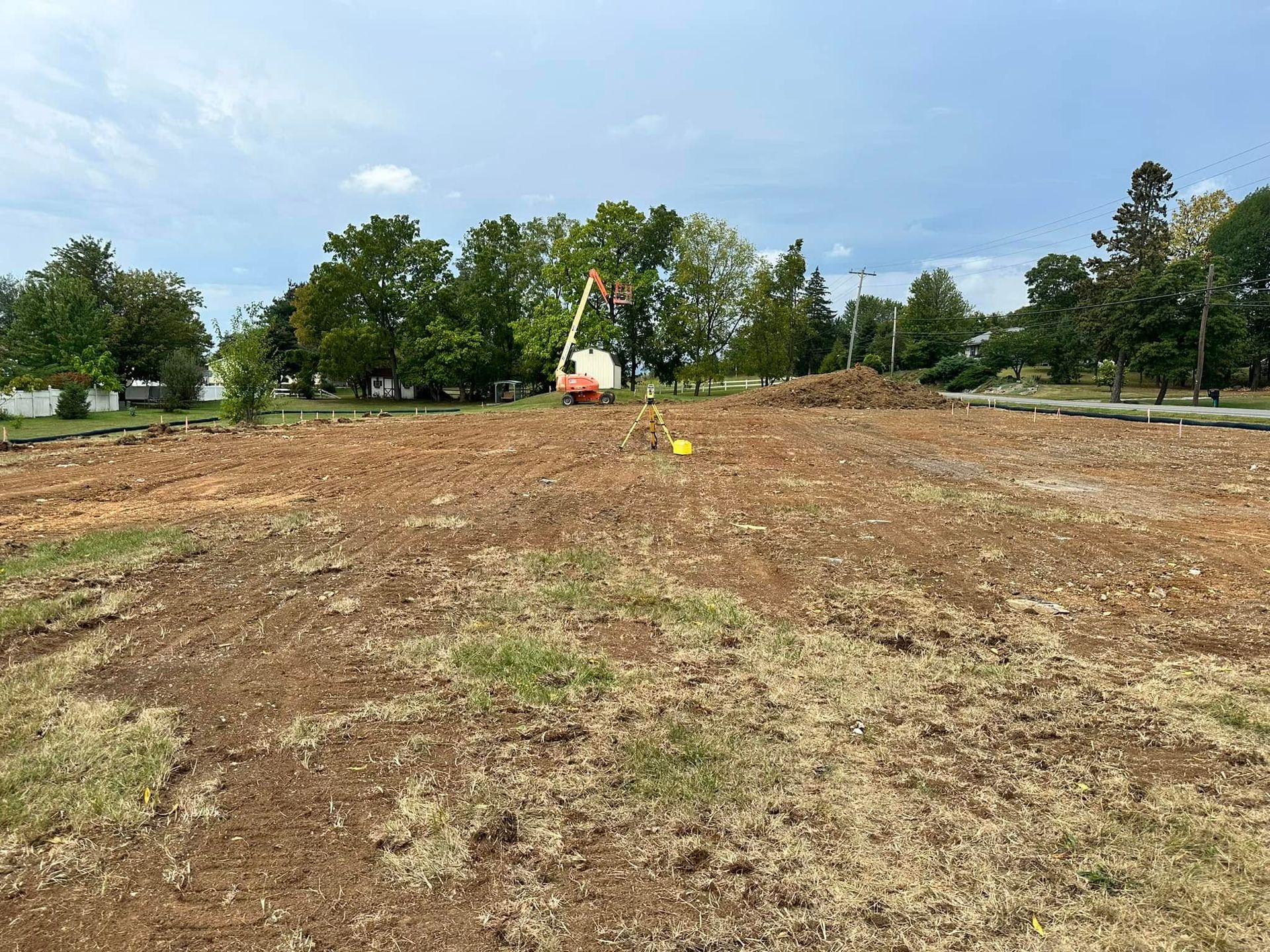 A cleared dirt construction site with scattered dry grass, surrounded by trees under a blue, cloudy sky.