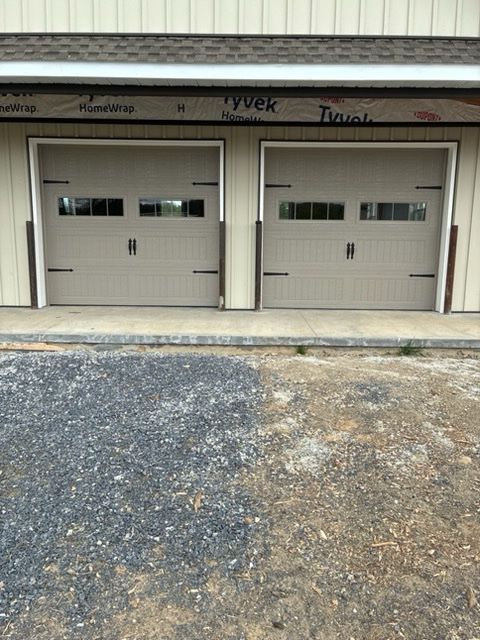 Two beige garage doors with small windows under a metal awning, beside a gravel drive.