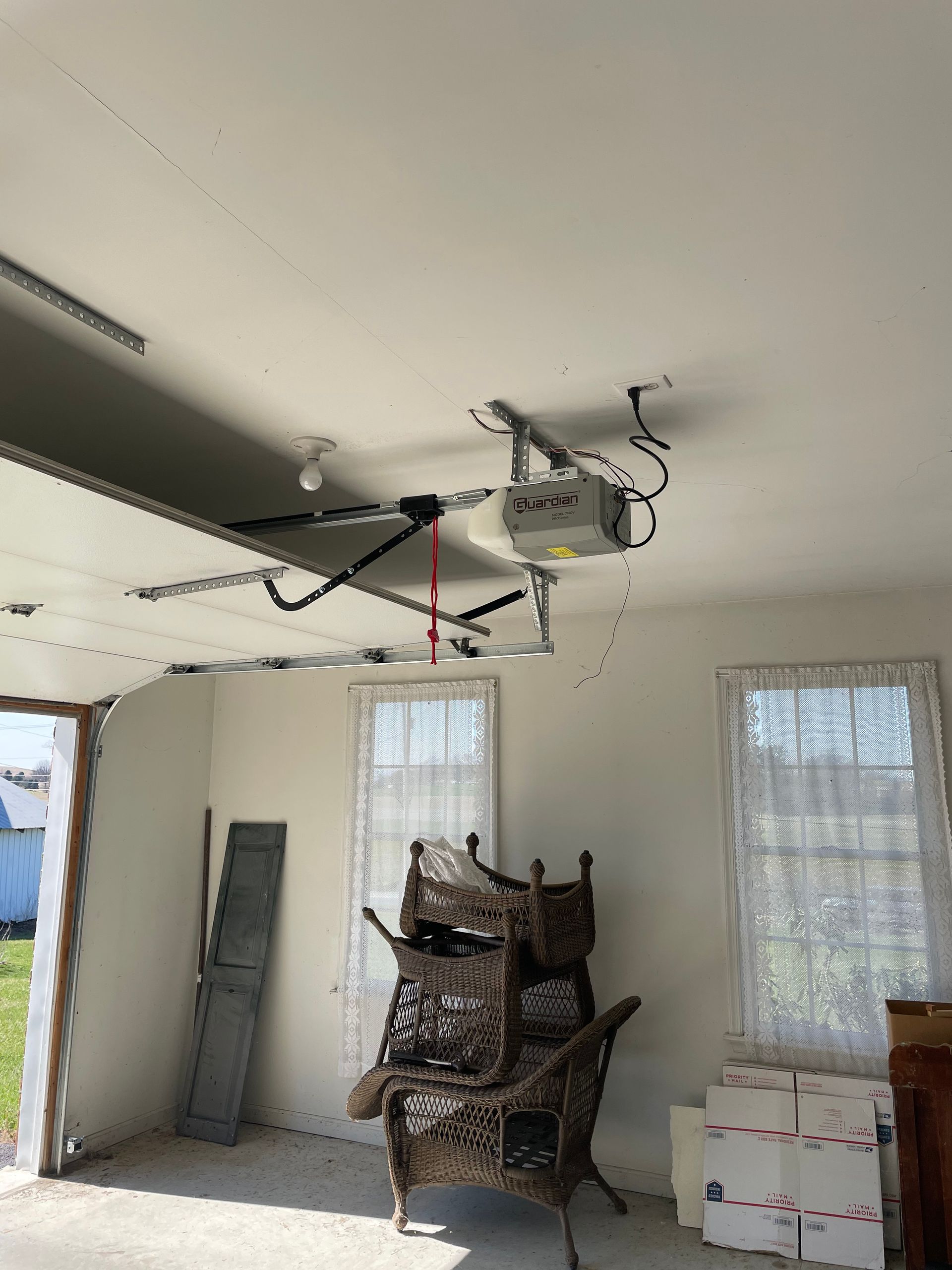 A garage interior features a ceiling-mounted door opener and a stack of three wicker chairs near two windows.
