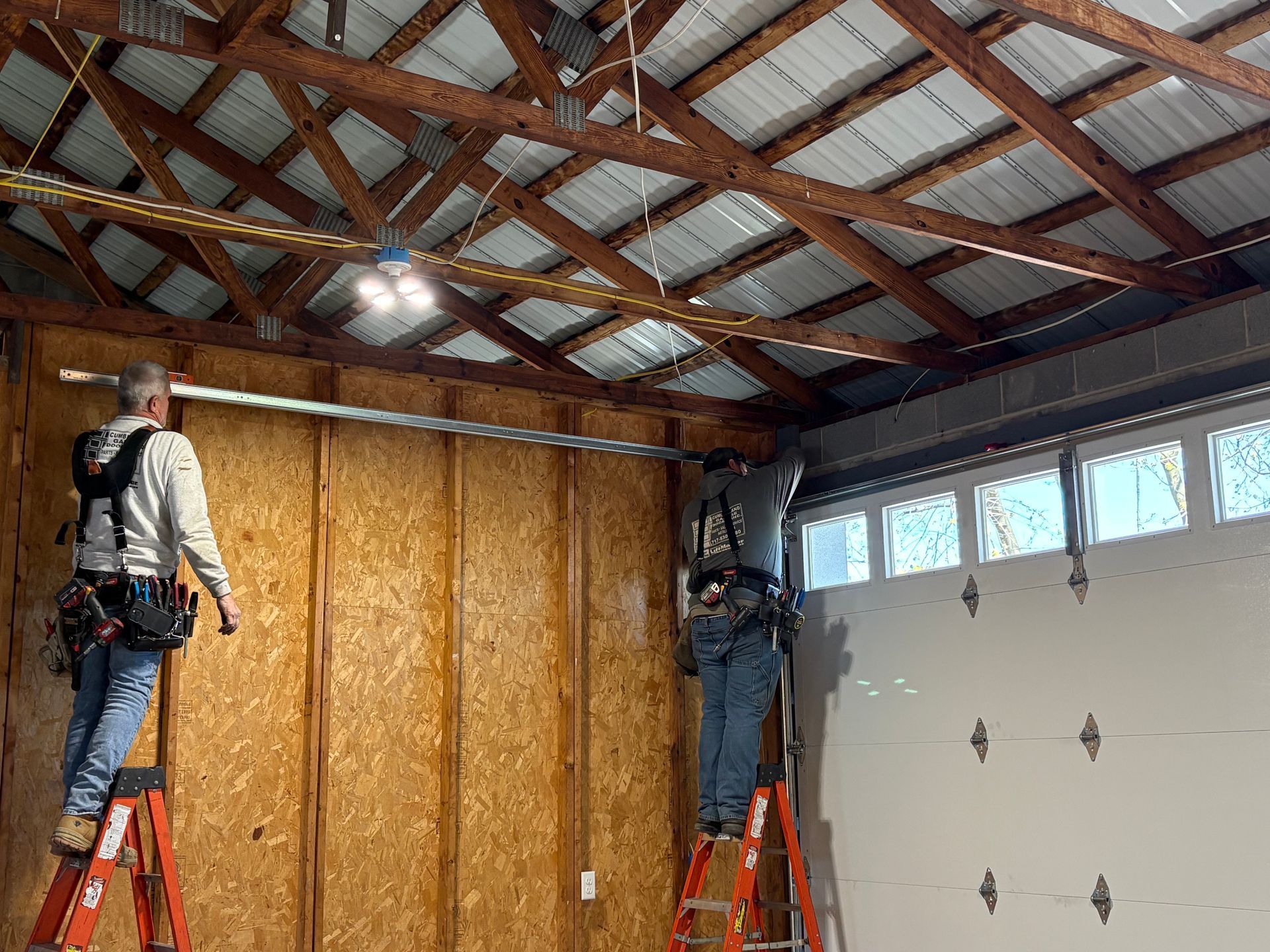 Two workers on stepladders install a metal track above a white garage door inside a wooden-walled garage.