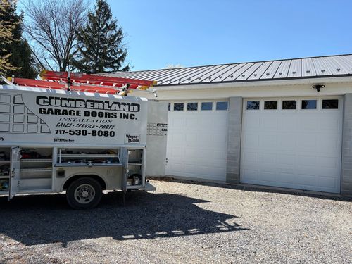 A dark gray garage door with windows on a house with stone-patterned siding and a nearby garden.