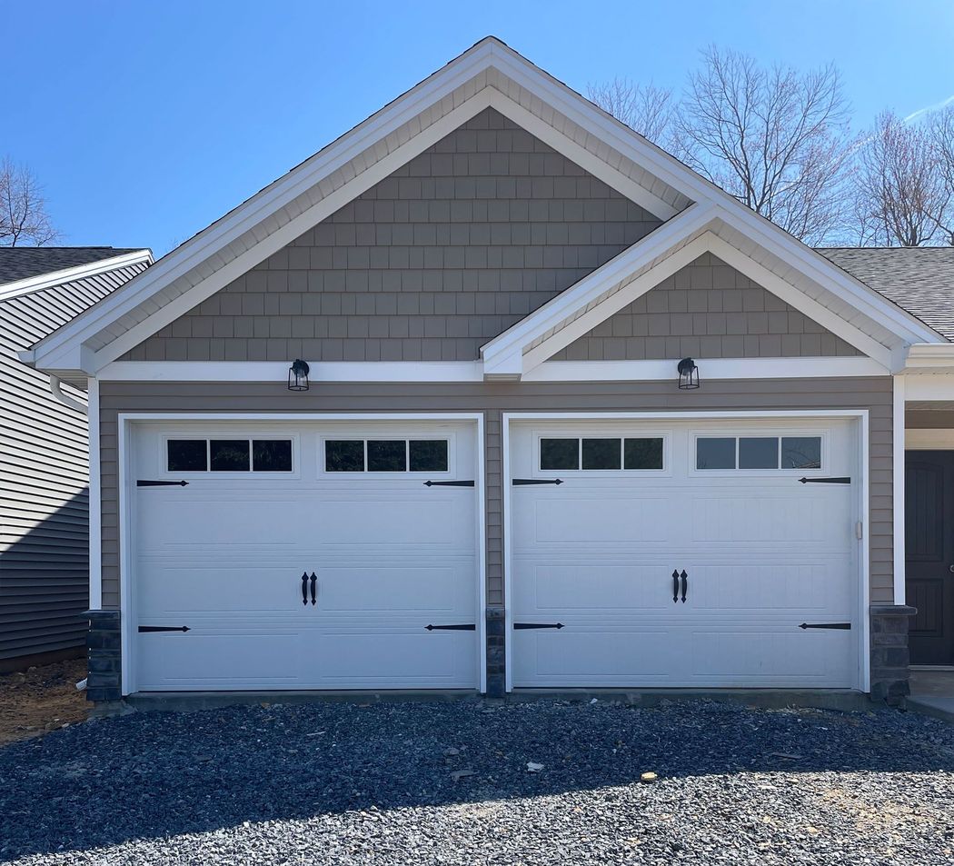 A two-car garage with white doors, black hinges, and light-brown siding under a blue sky on a gravel driveway.