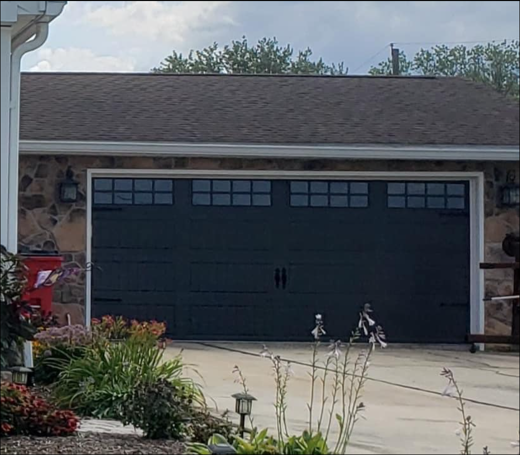 A dark gray garage door with windows on a house with stone-patterned siding and a nearby garden.