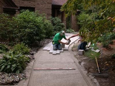 Two men are working on a sidewalk in front of a house.