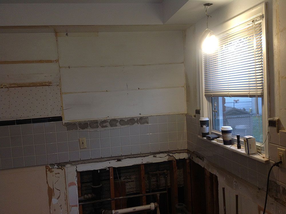 Kitchen under renovation; exposed studs, partially removed tile, and insulation; window with blinds.