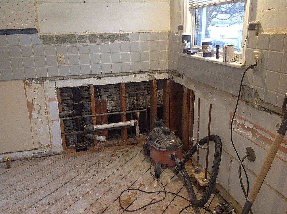 Kitchen undergoing renovation, with exposed plumbing, wood framing, and tile walls.