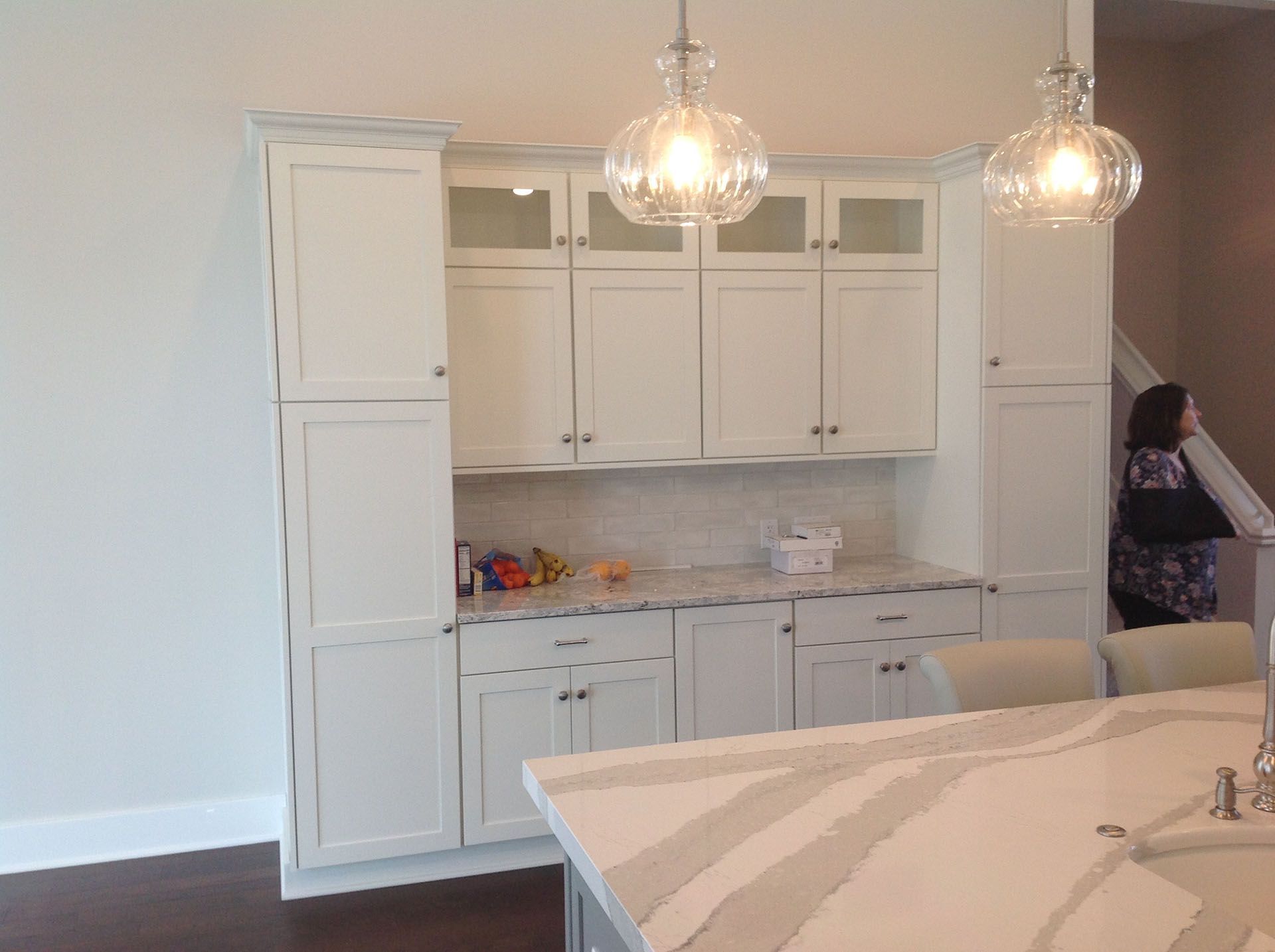 White kitchen cabinets with glass pendant lights, and a woman near stairs.