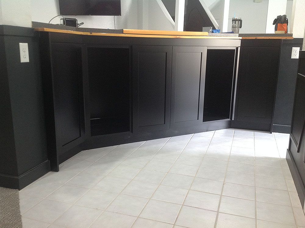 Black bar cabinets under a wooden countertop, set on white tile flooring. Electrical outlets on the walls.