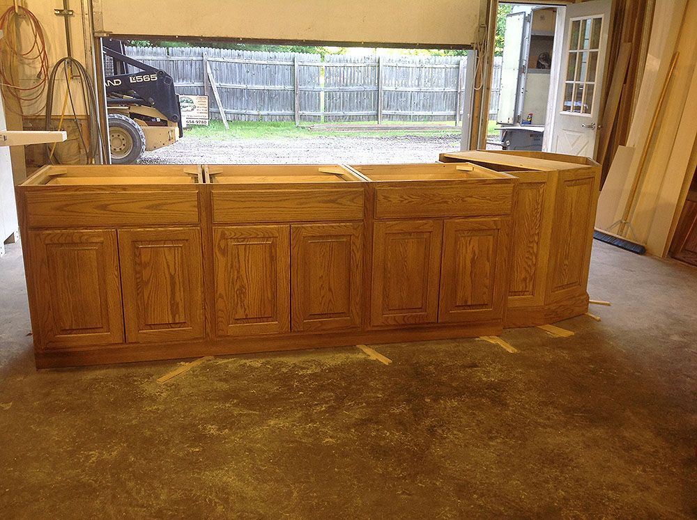 Wooden kitchen cabinets in a workshop, partially assembled. Sawdust on the floor, open garage door behind.