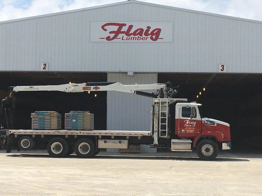 A flatbed truck is parked in front of a building that says flaig