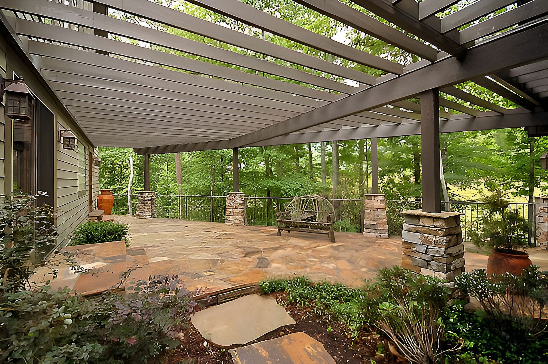 A stone patio under a curved wooden pergola, overlooking a lush green forest, with a rustic stone pillar in the foreground.