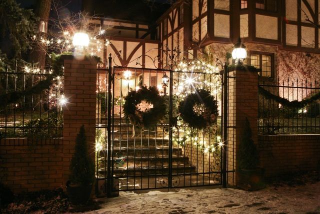 Festive house entrance with lit wreaths, brick gate, and snow at night