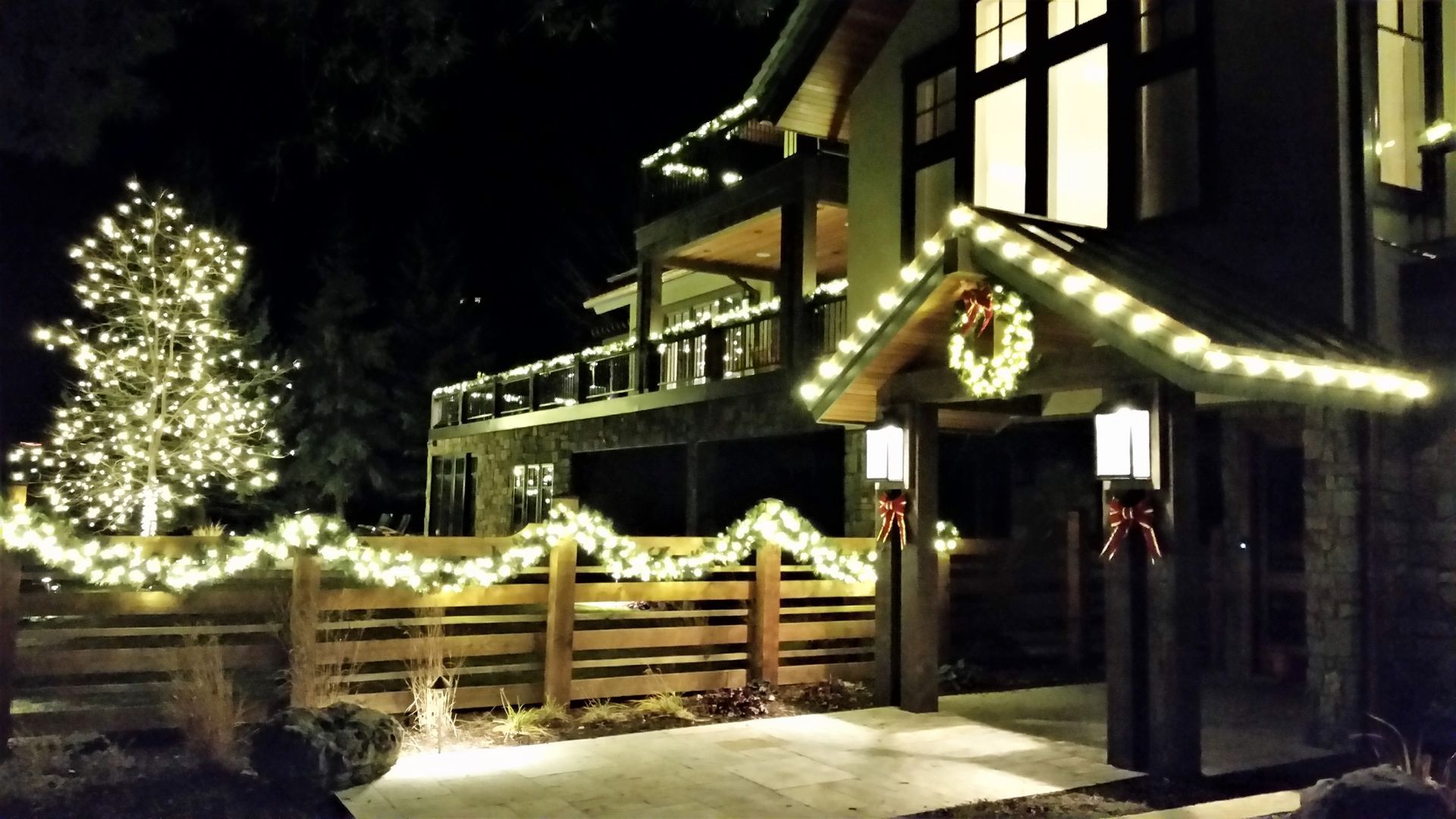 Snowy house decorated with holiday lights, lit tree, and wooden fence at night