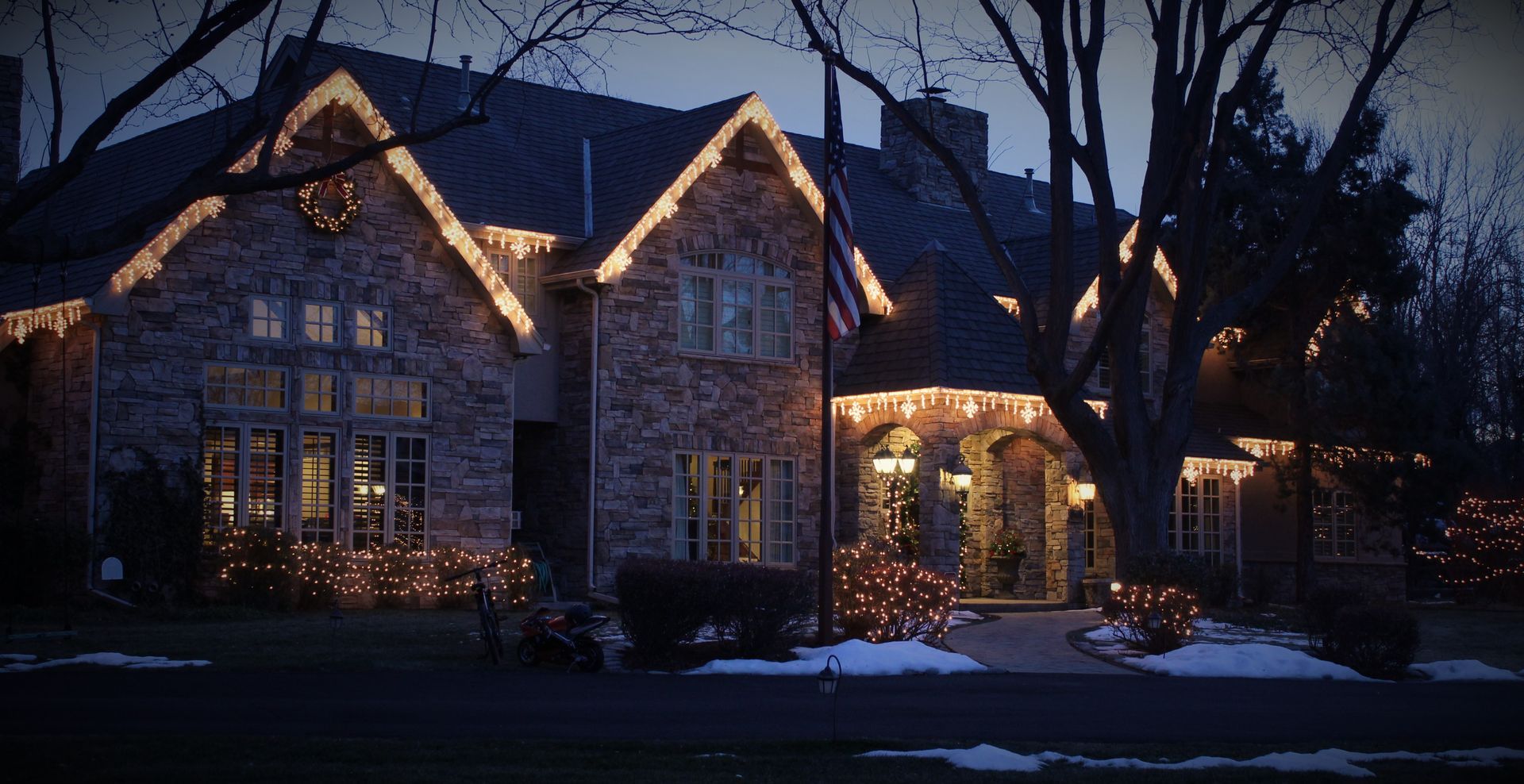 Snowy stone house at dusk with warm lights glowing from windows and entryway