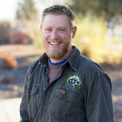Man with curly brown hair and beard smiles outdoors, wearing a gray jacket with a patch.