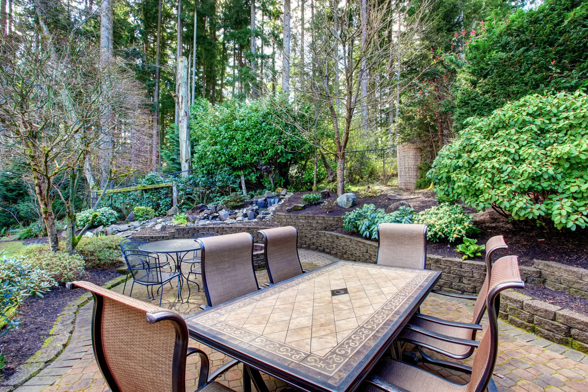 A tile-topped dining table with chairs sits on a stone patio in a lush, wooded backyard garden.