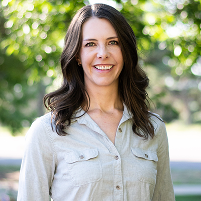 Woman smiles, wearing a light blue button-down shirt, standing outdoors in front of leafy trees.