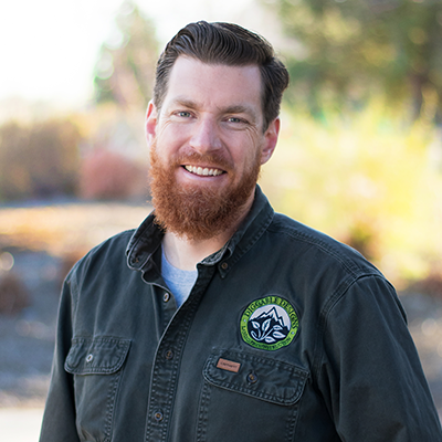 Man with red beard smiles, wearing a green work shirt with a mountain logo; outdoors.